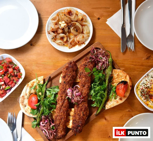 Bir tepsi üzerinde servis edilen, közlenmiş biber ve domates, sumaklı soğan ve taze maydanozla süslenmiş üç adet iştah açıcı Adana kebap, yanında gavurdağı salatası, tereyağlı soğan ve baharatlı yoğurtlu bir meze ile İstanbul'un en iyi et restoranları deneyimini yansıtan zengin bir kebap sofrası.