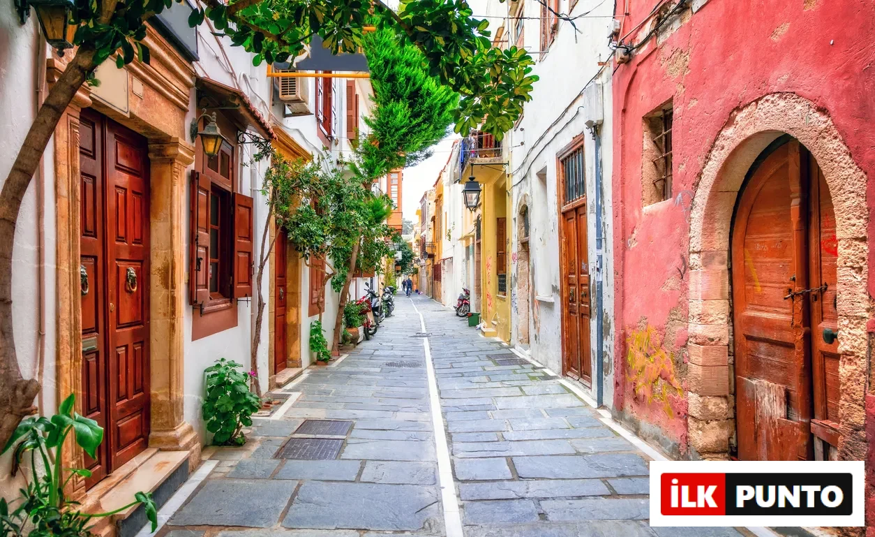 Yemyeşil ağaçlar ve bitkilerle bezenmiş, beyaz, sarı, turuncu ve pastel pembe tonlarında eski binaların sıralandığı, ahşap kapı ve pencereleriyle dikkat çeken taş döşeli dar bir Girit sokağı, uzaklarda yürüyen insan silüetleriyle Akdeniz atmosferini yansıtıyor.