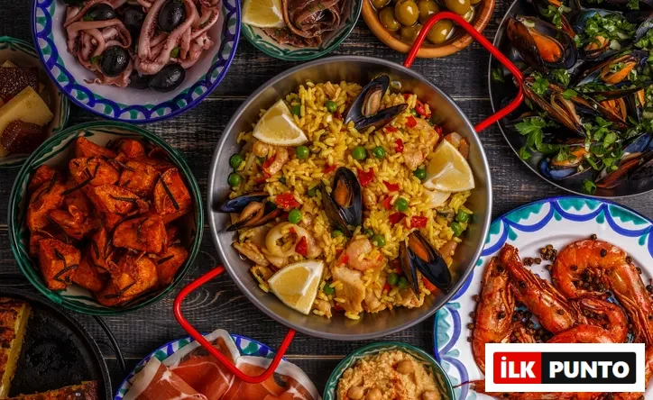 Ahşap bir masa üzerinde, ortada midye, karides, kalamar, tavuk, bezelye ve limon dilimleriyle süslenmiş büyük bir paella tavası, çevresinde ızgara karidesler, maydanozlu midyeler, ahtapot salatası, kızarmış patatesler, dilimlenmiş İspanyol omleti, jambon, ançuez ve zeytin gibi çok sayıda renkli ve iştah açıcı İspanyol tapas çeşidiyle dolu zengin bir İspanyol mutfağı ziyafeti sunuluyor.