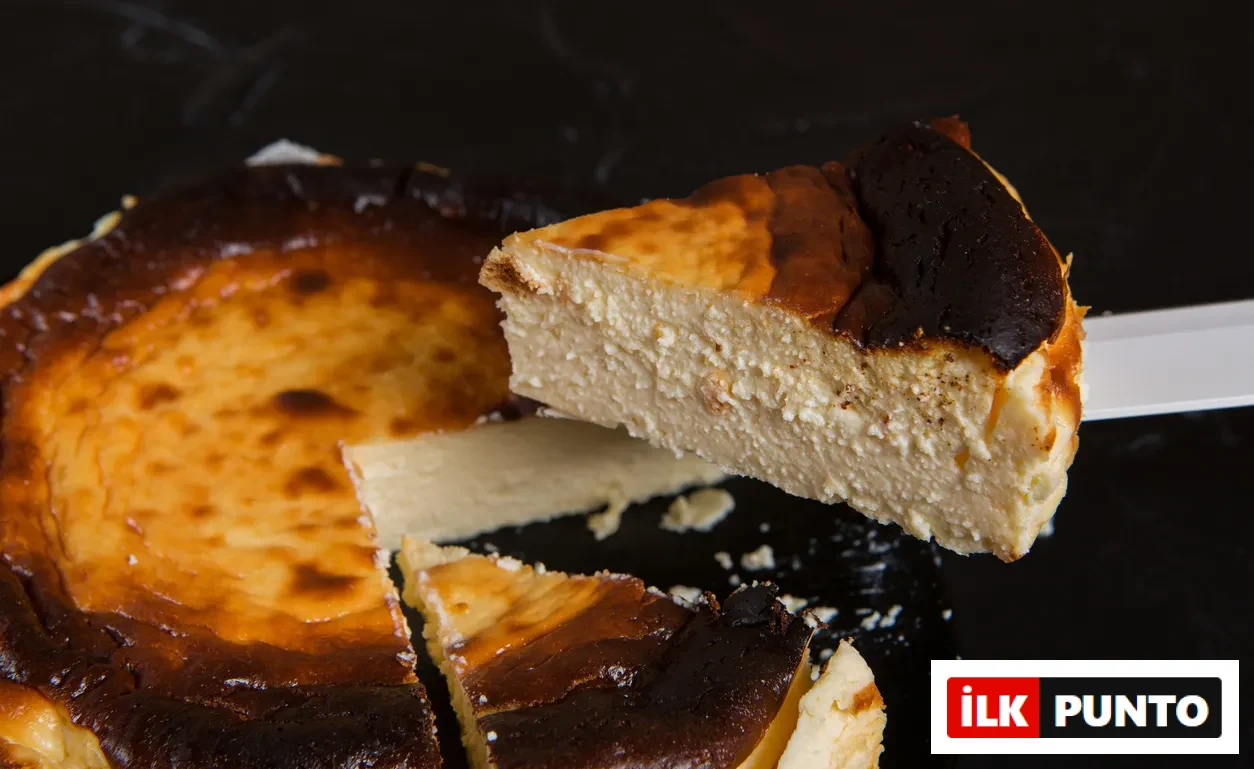 Koyu bir zeminde duran, üzeri koyu karamelize ve hafif yanık kabuklu, içi ise yoğun ve kremsi bir dokuya sahip bütün bir San Sebastian cheesecake'ten beyaz bir spatula yardımıyla bir dilim nazikçe servis ediliyor, yanında diğer kesilmiş dilimler de görülebiliyor.
