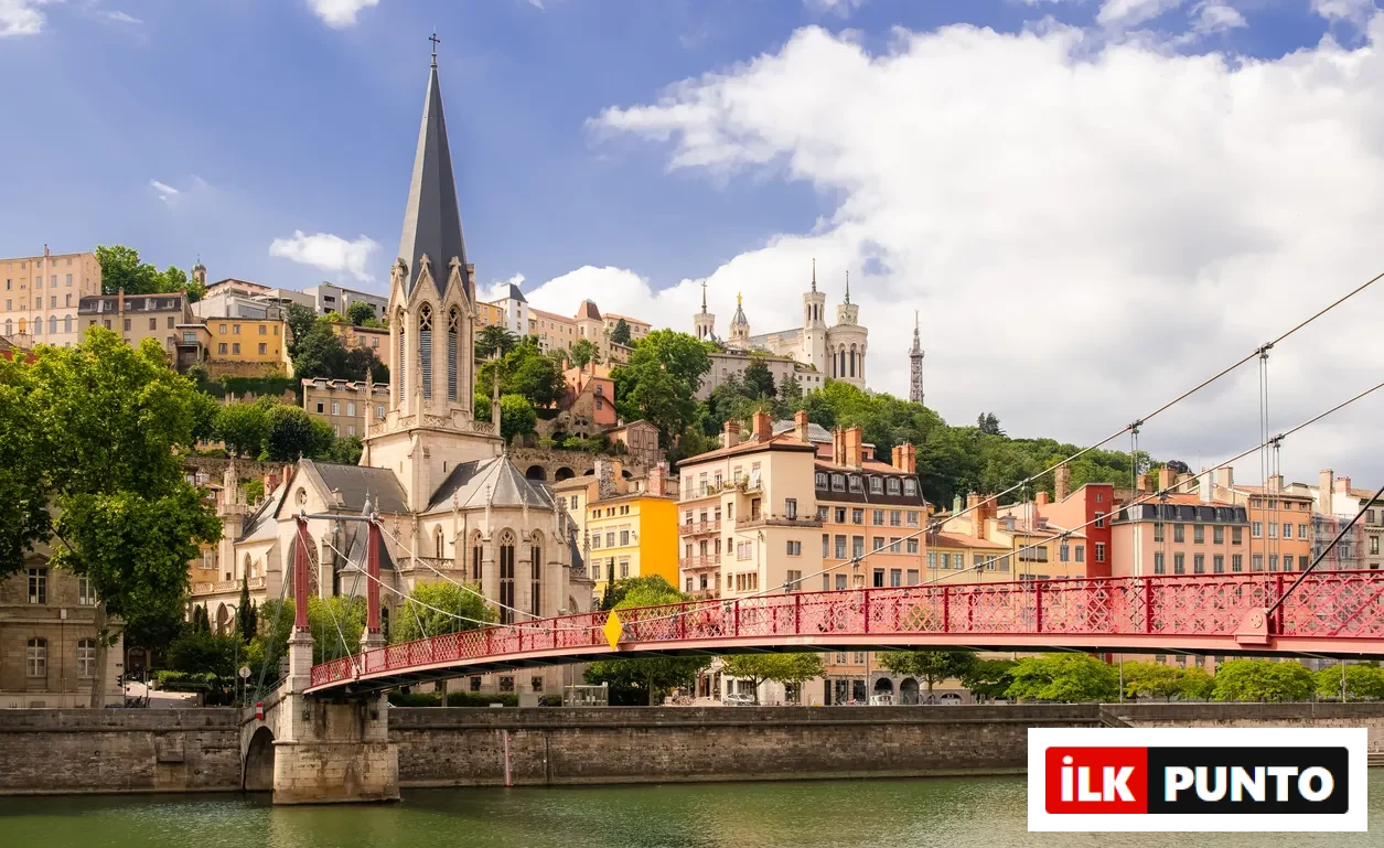 Lyon'da, yeşil Saône Nehri üzerinde kemerli, kırmızı bir asma köprü, solunda Sainte-Georges Kilisesi'nin sivri gotik kulesi, yamaçta sıralanmış tarihi renkli binalar ve en tepede ikonik Fourvière Bazilikası'nın göz kamaştırıcı mimarisi bir arada görülüyor.