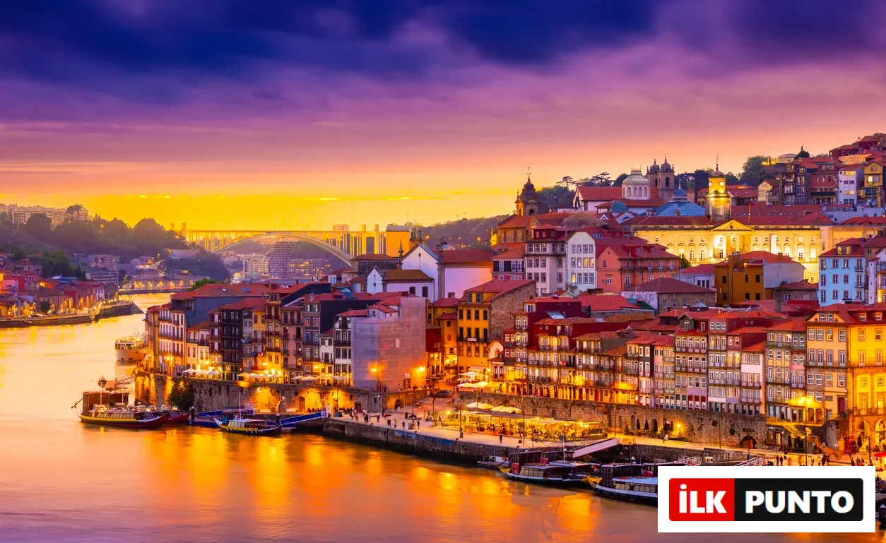 Porto şehrinin Douro Nehri boyunca sıralanmış tarihi ve renkli binalarının, gün batımının canlı mor ve turuncu tonlarıyla gökyüzünü boyadığı, su üzerinde parıldayan ışıkların ve hareketli rıhtımın büyüleyici bir şehir manzarası sunduğu geniş açılı bir görüntü.