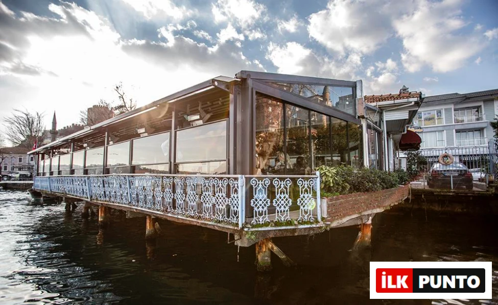 Modern cam duvarları ve açılır kapanır tavanıyla su üzerinde ahşap direkler üzerine inşa edilmiş, uzun ve zarif bir restoran, sol arka planda Rumeli Hisarı'nın tarihi siluetini barındırırken, bulutlu gökyüzünden süzülen güneş ışıklarıyla pırıl pırıl parlıyor.