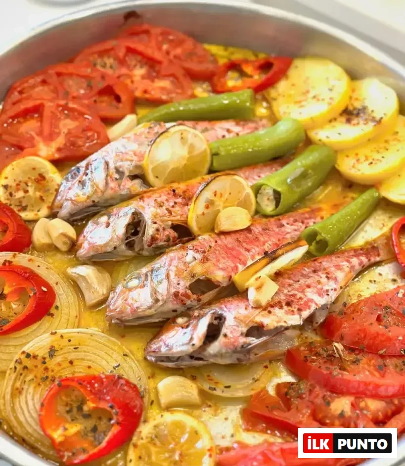 Büyük, parlak metal bir tepside, zeytinyağında pişmiş ve üzerine baharatlar serpilmiş, limon dilimleri ve sarımsak taneleri ile süslenmiş barbun balıkları; çevresine düzenli şekilde yerleştirilmiş taze domates, patates, halka soğan, yeşil ve kırmızı biber dilimleriyle görsel olarak zengin bir deniz mahsulü yemeği sunuluyor.