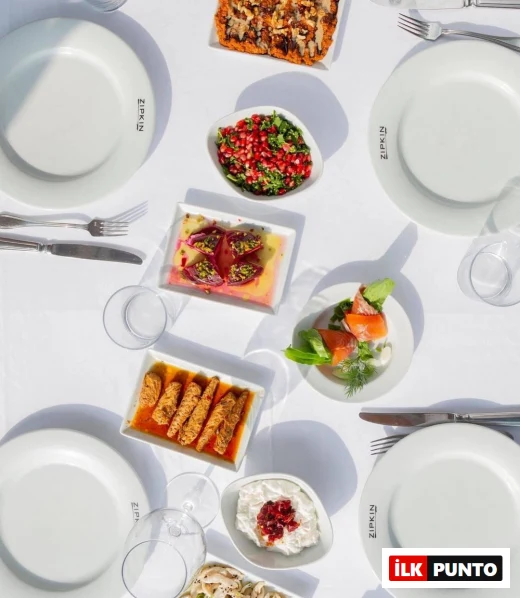 Beyaz örtülü bir masada yukarıdan çekilmiş bu görüntüde, ZİPKİN logolu boş tabaklar, nar taneli yeşil salata, somon füme ruloları, mantar salatası, kırmızı soslu kızarmış deniz mahsulleri ve fıstıklı pembe mezeler dahil zengin çeşitlilikte Ege ve Akdeniz mutfağına özgü başlangıçlar sergileniyor.