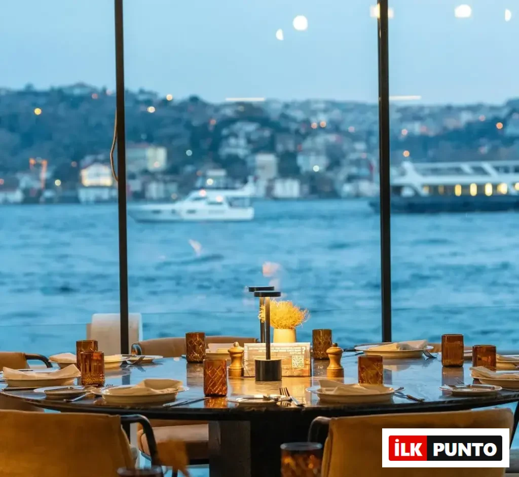 Boğaz manzaralı bir restoranda, şık bir masa düzeniyle akşam yemeği için hazırlanmış yuvarlak masa, suya yansıyan şehir ışıkları ve yakından geçen tekneler eşliğinde davetkar bir atmosfer sunuyor.