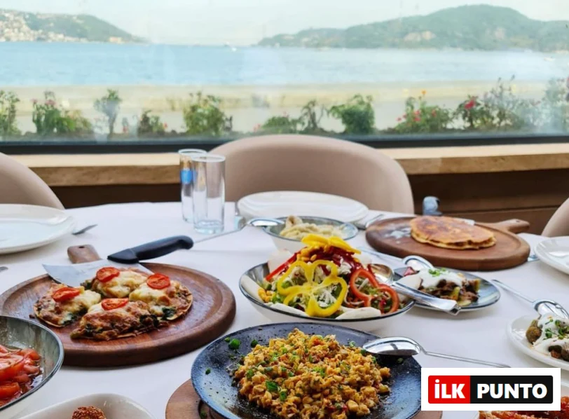 Deniz kenarında, huzurlu bir ortamda kurulan beyaz örtülü bir masada, peynirli ve domatesli fırın yemeği, rengarenk biberli salata, tavada çırpılmış yumurta, krepler ve çeşitli mezelerle dolu zengin bir öğün sergileniyor.
