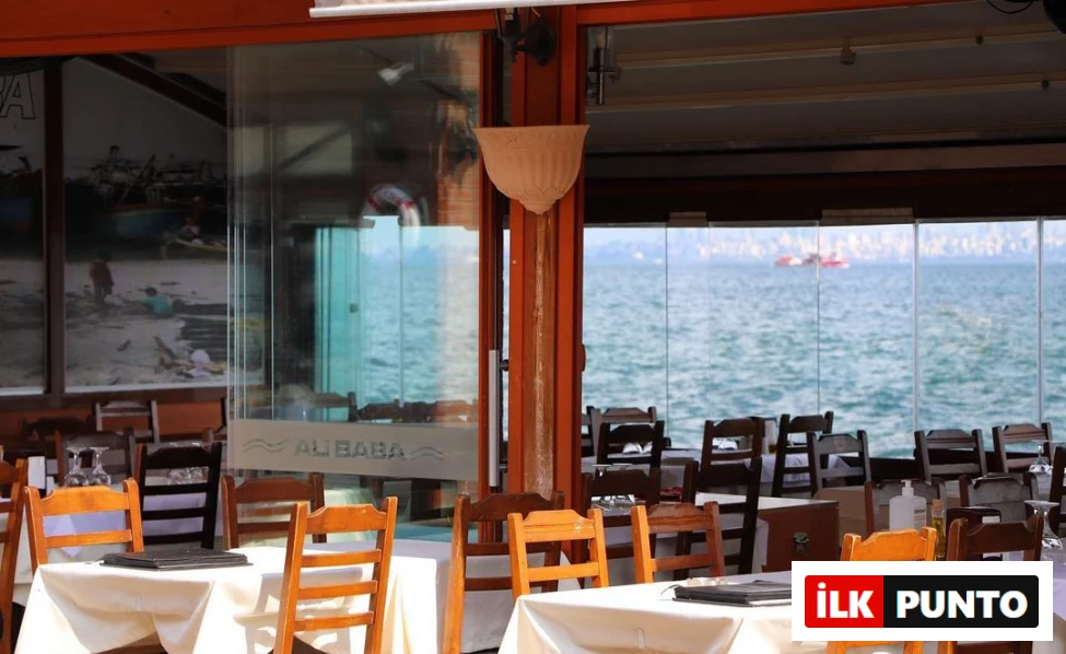 Büyükada'da yer alan Ali Baba restoranının iç mekanından çekilen bu fotoğrafta, deniz manzaralı camların önünde boş bekleyen beyaz örtülü masalar ve ahşap sandalyeler, uzaktaki sakin denizi ve bir teknenin siluetini gözler önüne seriyor.
