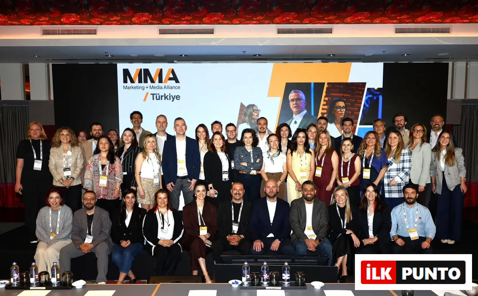MMA Marketing + Medya İttifakı Türkiye logosunun büyük bir ekranda belirgin şekilde görüldüğü bir konferans salonunda, farklı yaş ve cinsiyetten pazarlama ve medya profesyonelleri, etkinliğin ruhunu yansıtan kalabalık bir grup fotoğrafı için bir araya gelmiş.