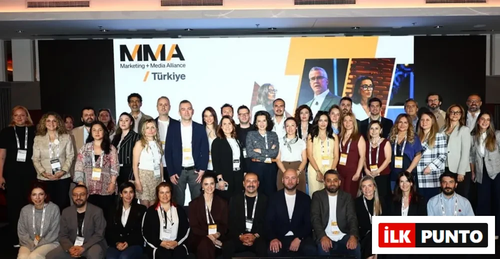MMA Marketing + Media Alliance Türkiye'nin çeşitli kademelerinden yöneticiler ve profesyonellerin, markanın logosunun ve geçmiş etkinlik görsellerinin yer aldığı büyük bir ekran önünde topluca poz verdiği, katılımcıların yaka kartlarıyla kurumsal kimliği yansıttığı canlı bir etkinlik fotoğrafı.