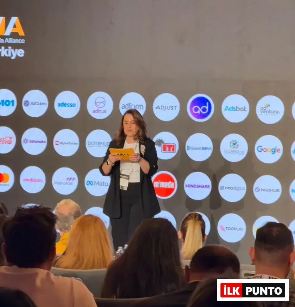 Sahne üzerinde, arkasında birçok şirketin logosu ve MMA Türkiye yazısı bulunan bir fonda, Didem Namver siyah blazer ceketi ve beyaz bluzuyla elinde sarı not kağıtları tutarak dikkatle sunumunu yapıyor, ön planda ise dinleyicilerin başları görülüyor.
