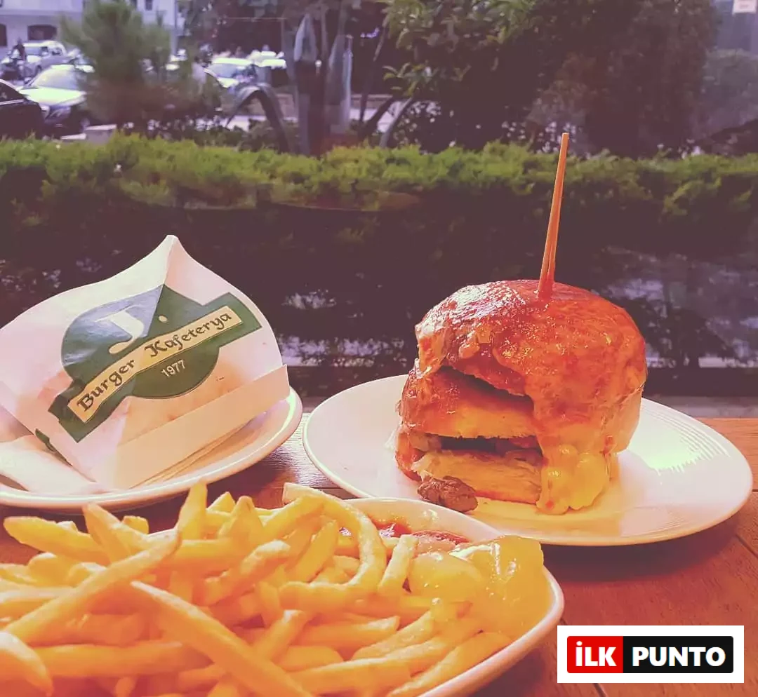 Duru Akgül'ün şeflerin lezzet rotaları kapsamında favori hamburger mekanlarından J Burger'da çekildiği anlaşılan bu yakın planda, erimiş peynirleri akan büyük bir hamburgerin yanında bol porsiyon patates kızartması ve üzerinde Burger Kafeterya 1977 yazan bir paket ahşap masada iştah açıcı duruyor.