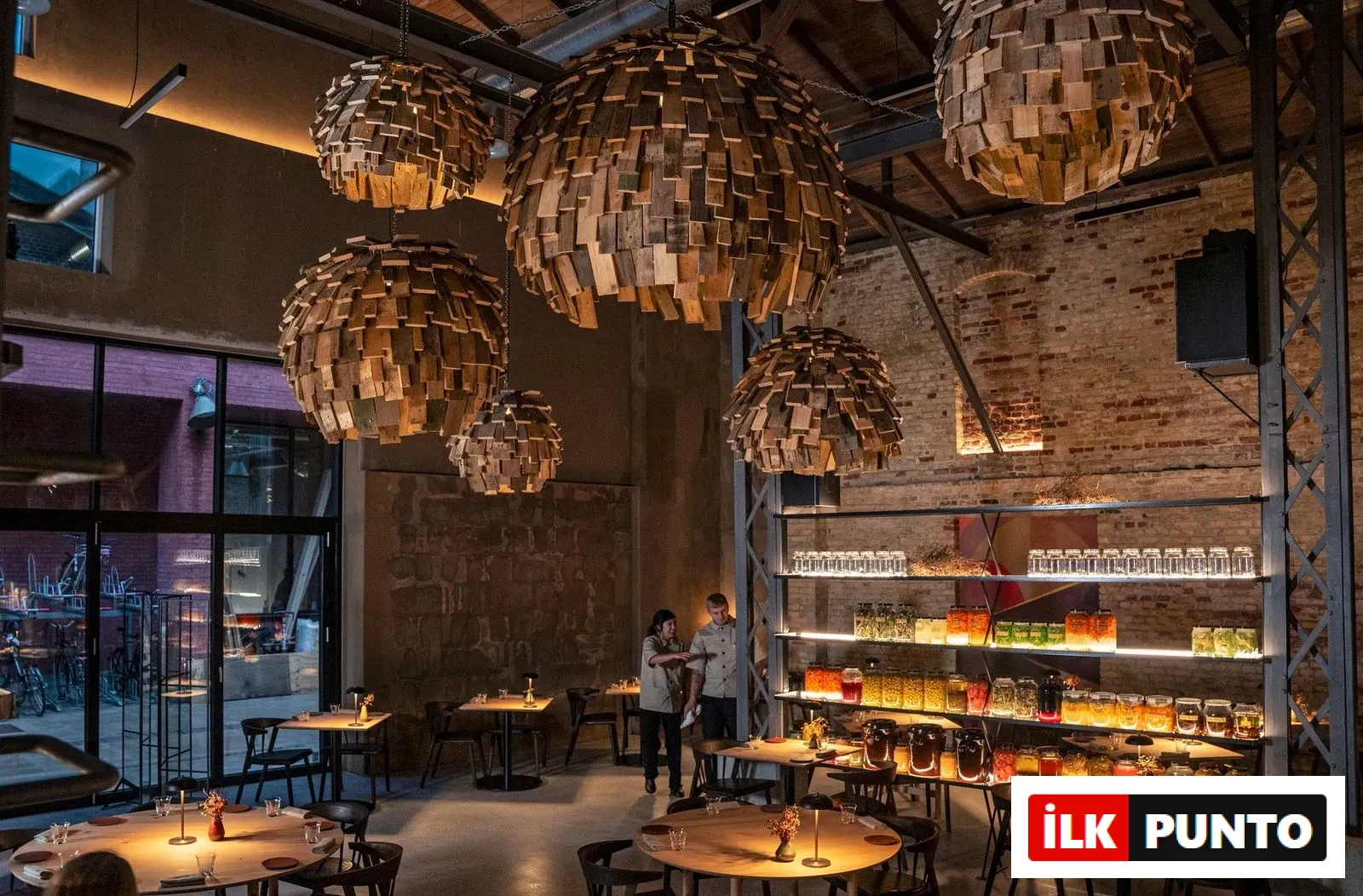Esse Kopenhag restoranının iç mekanında, ahşap parçalarından yapılmış büyük, kubbe şeklinde avizeler tavandan sarkarken, duvarlardaki raflarda rengarenk fermente edilmiş gıdalar ve turşularla dolu cam kavanozlar ve sıralanmış bardaklar parlak bir şekilde sergileniyor; loş ışıklı masalarda oturma düzeni ve iki çalışan görünmekte.