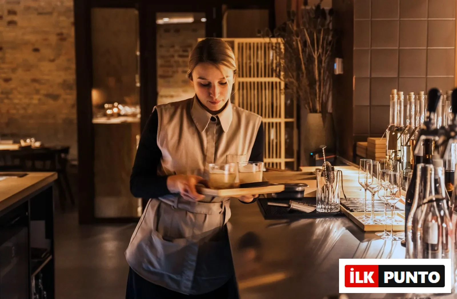 Kopenhag'daki zarif Esse restoranının loş ışıklı, sıcak atmosferinde, yelekli önlüklü genç bir kadın garson, paslanmaz çelik bar tezgahının önünde, içinde açık renkli içecekler bulunan iki bardağı ahşap bir tepside dikkatle taşıyor ve servis etmeye hazırlanıyor.
