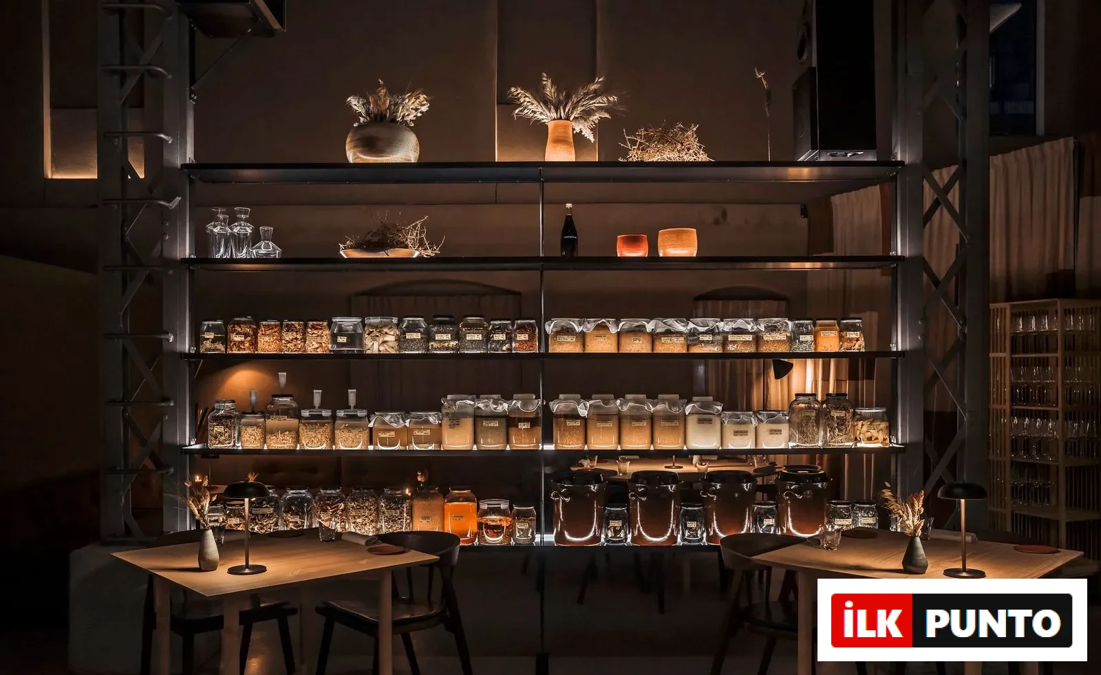 Kopenhag'daki Esse restoranının loş ışıklı iç mekanında, endüstriyel raflarda sıralanmış, içinde çeşitli tahıllar, baharatlar ve sıvıların bulunduğu yüzlerce cam kavanoz dikkat çekiyor; ön planda ise sıcak bir atmosfer sunan iki ahşap masa ve sandalye takımı yer alıyor.