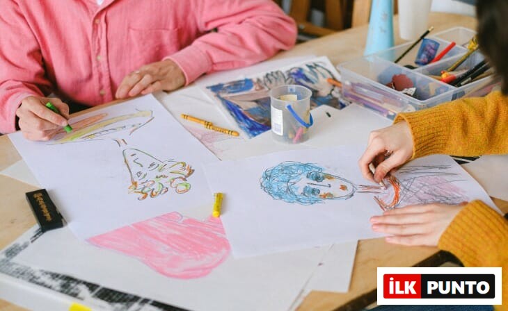 Ahşap bir masada oturan iki kişinin, biri pembe diğeri hardal sarısı kazaklı, farklı renklerde pastel boyalarla yüz çizimleri ve soyut şekillerin olduğu beyaz kağıtları boyarken, evden çalışırken yaratıcılığı ve motivasyonu artıracak mola ve ilham kaynakları bulmanın önemini vurgulayan samimi bir an.