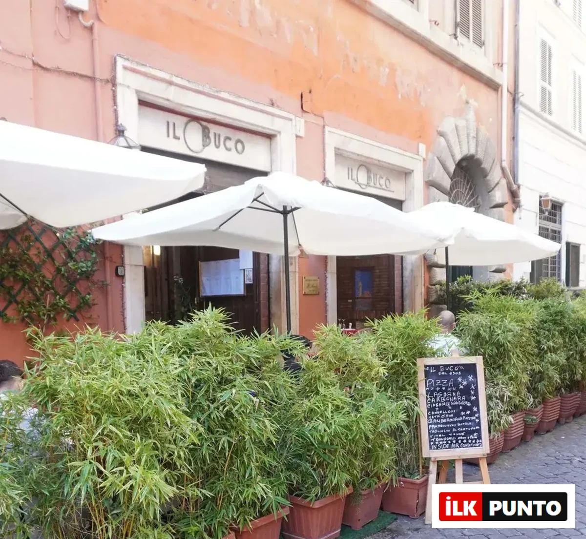 Romantik bir Avrupa sokağında yer alan, ön cephesi turuncu renkli IL BUCO adlı ünlü restoranın, beyaz güneş şemsiyeleri, saksılarda sıralanmış gür yapraklı bitkiler ve kaldırımda duran bir menü tahtasıyla temasına uygun dış cephesi.