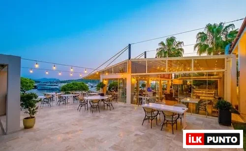 Akşamüstü maviliğinde, parlak ışıklarla aydınlatılmış ferah mermer terasta, denize nazır modern cam cepheli restoranın önünde, uzakta demirli teknelerin ve yemyeşil kıyıların görüldüğü, şık masa ve sandalyelerin misafirlerini beklediği keyifli bir açık hava mekanı.