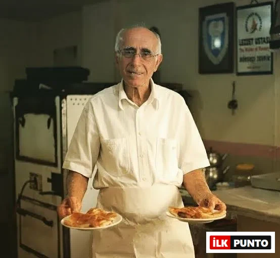 Beyaz gömlek ve önlük giymiş, gözlüklü ve güler yüzlü Tevfik Usta, arka planda geleneksel fırın ve Lezzet Ustası plaketinin asılı olduğu mutfak ortamında, her iki elinde de yeni pişmiş, altın rengi böreklerle dolu tabakları tutarak kameraya gülümsüyor.