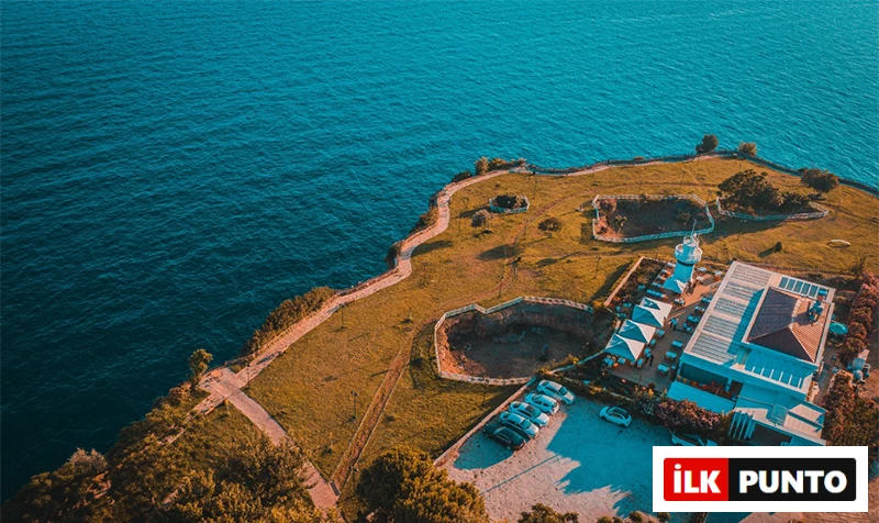 Antalya'nın en iyi restoranlarından Fener Bababurnu, turkuaz Akdeniz'in kıyısındaki yemyeşil bir falez üzerinde, açık hava oturma alanları, beyaz bir deniz feneri ve otoparkı ile çevrili modern binalarıyla kuşbakışı görünüyor, ziyaretçilerine eşsiz bir manzara sunuyor.