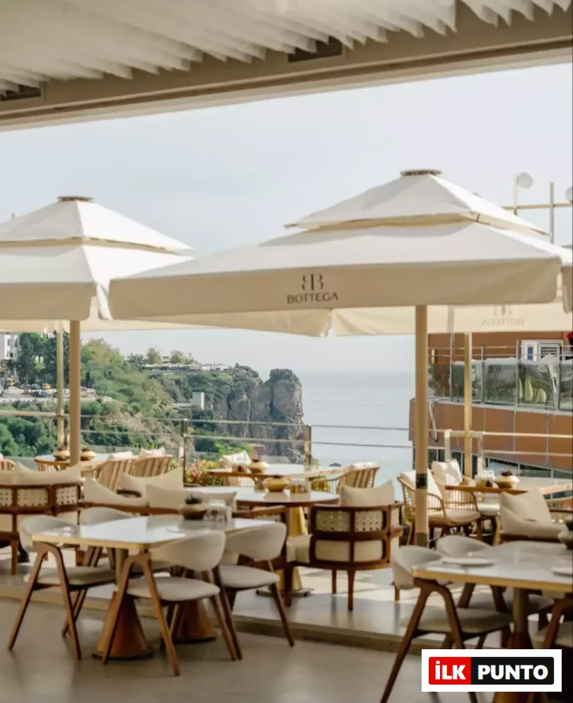 Masmavi denize ve etkileyici kayalıklara bakan, Bottega logolu bej şemsiyelerin gölgelediği ferah bir açık hava restoran terası, modern ahşap detaylı beyaz masa ve sandalyelerle misafirlerini ağırlamaya hazır, huzurlu bir atmosfer sunuyor.