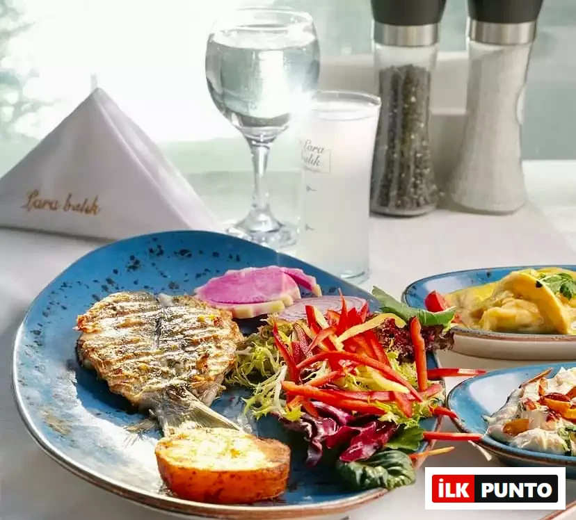 Masa üzerinde, mavi bir tabakta ızgara edilmiş taze balık, yanında rengarenk Akdeniz salatası, pembe turp dilimleri ve fırınlanmış patates ile sunulurken, arka planda Lara Balık yazılı rakı kadehi ve baharatlıklar, Antalya'nın zengin deniz ürünleri ve Akdeniz mutfağının eşsiz lezzetlerini deneyimlemek için ideal bir gastronomi anını yansıtan bir sofrayı tamamlıyor.