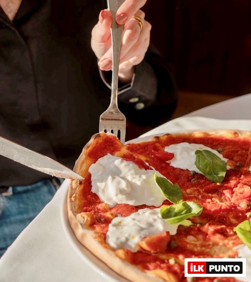 Etiler'deki Da Mario gibi seçkin bir mekanda, güneş ışığı alan beyaz masa üzerinde taze burrata peyniri, domates sosu ve fesleğen yapraklarıyla süslenmiş İtalyan pizzasının, ojeli bir el tarafından çatal ve bıçakla kesilerek servis edilmeye hazırlandığı iştah açıcı bir yakın plan.