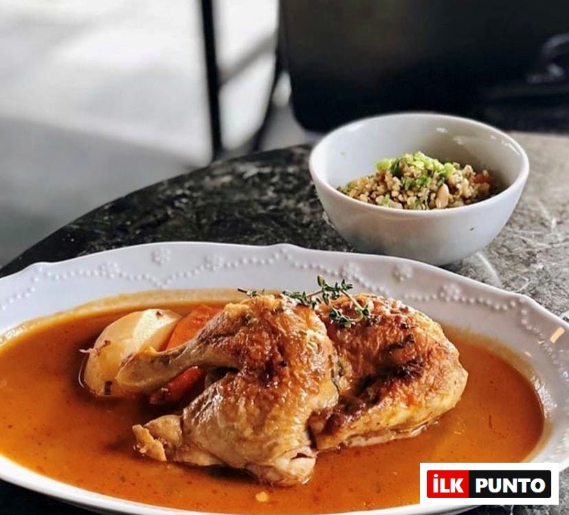 Altın rengi kızarmış tavuk parçaları, patates ve havuç dilimleriyle birlikte zengin turuncu bir sos içinde, kenarları oymalı oval beyaz bir tabakta iştah açıcı bir şekilde sunuluyor; yanında ise taze yeşilliklerle süslenmiş tahıl salatası bulunan küçük beyaz bir kase Etiler'deki en iyi mekanlardan Delicatessen'in lezzetlerini yansıtıyor.