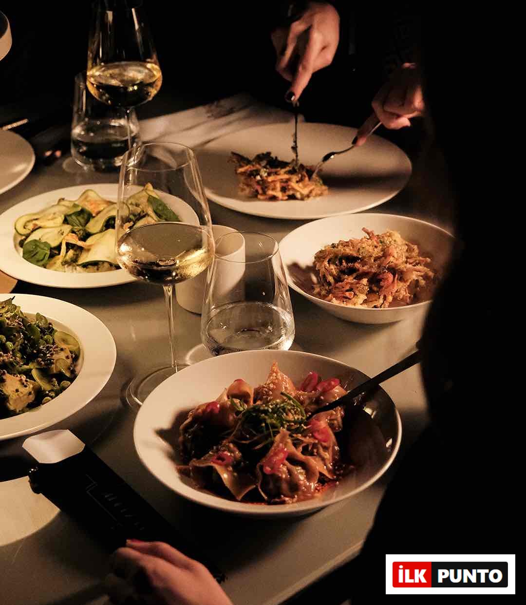 Loş ışıklandırılmış bir restoranda veya yemek masasında, beyaz tabaklarda sunulmuş çeşitli yemekler (makarna, soslu bir ana yemek ve salata gibi), şarap kadehleri ve su bardaklarıyla dolu samimi bir akşam yemeği anı; bir misafir yemeğini çatal bıçak kullanarak yiyor.