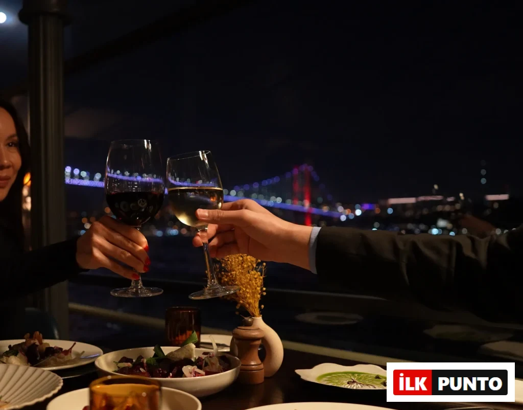 Gece karanlığında, Boğaz Köprüsü ve şehir ışıklarının parladığı muhteşem bir manzaraya karşı, özenle hazırlanmış bir yemek masasında kırmızı ve beyaz şarap kadehleriyle kadeh kaldıran iki kişi, şık ve kutlama dolu bir akşam yemeği anını yansıtıyor.