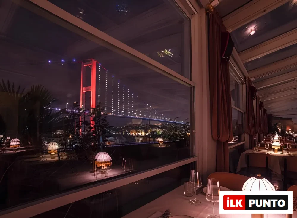Gecenin karanlığında kırmızı ışıklarla aydınlatılmış Boğaz Köprüsü manzarasının büyük pencerelerden göründüğü şık bir restoranda, özenle hazırlanmış masalarda kadehler ve etrafa sıcak loş ışık yayan masa lambalarıyla samimi bir akşam yemeği atmosferi.