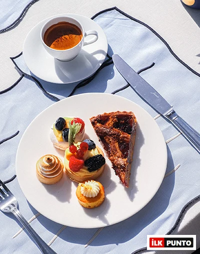 Parlak güneş ışığı altında, beyaz bir tabakta frambuaz, böğürtlen ve kivi dilimleriyle süslenmiş üç küçük meyveli tart, spiral şekilli bir beze, üzeri kremalı diğer bir mini pasta ve koyu renkli bir tart diliminin iştah açıcı bir şekilde sunulduğu, yanında metal çatal bıçak takımı, dalgalı desenli açık mavi masa örtüsü ve arka planda bir fincanın seçildiği zengin tatlı tabağı.