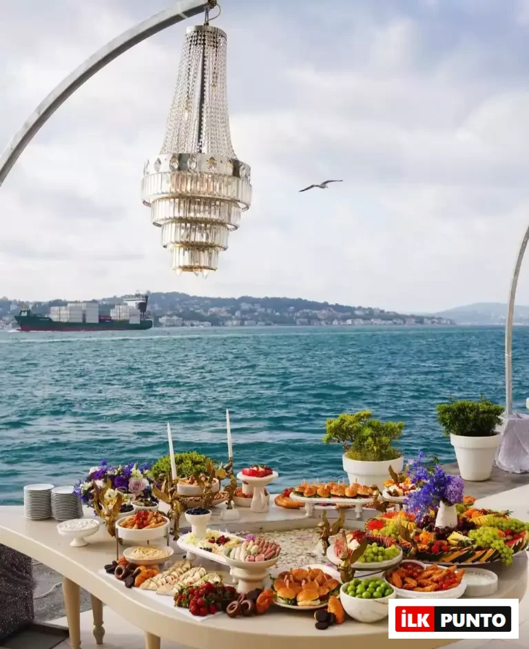 Ajia Hotel'in Boğaz manzaralı terasında, üzerinde şık kanepeler, meyveler ve çeşitli ikramlarla donatılmış uzun bir ziyafet masası, havada asılı duran kristal avize ve uzakta geçen gemilerle büyüleyici bir davet ortamı sergileniyor.