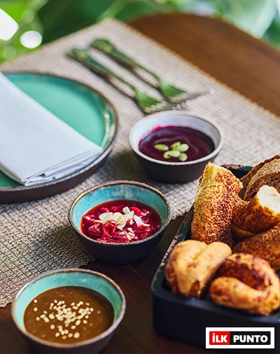 Ahşap bir masanın üzerinde sunulan zengin bir brunch masası detayı, içerisinde çeşitli altın rengi ekmekler ve çıtır hamur işlerinin bulunduğu siyah bir sepeti gösteriyor; yanında, yeşil kabak çekirdekleriyle süslenmiş mor renkli bir meyve sosu, kıyılmış bademlerle bezenmiş parlak kırmızı bir reçel ve beyaz susam taneleriyle zenginleştirilmiş kahverengi bir tahin veya fındık ezmesi olmak üzere üç farklı lezzetli dip sos küçük seramik kaselerde özenle yer alıyor.