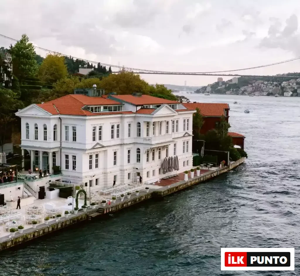 Boğaz'ın kıyısında konumlanmış, kırmızı kiremit çatılı, beyaz ve görkemli Ajia Hotel, su üzerindeki teraslarında şık açık hava etkinlik ve yemek düzenlemeleriyle dikkat çekerken, arka planda Boğaz Köprüsü ve karşı yaka manzarası uzanıyor.