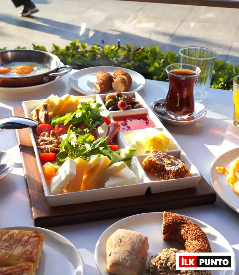 Güneşli bir ortamda beyaz bir masa üzerinde özenle hazırlanmış, peynir çeşitleri, zeytin, taze yeşillikler, reçel, bal peteği, sahanda yumurta, simit, börek ve çay gibi zengin içeriğe sahip, göz alıcı bir Türk kahvaltısı sergileniyor.