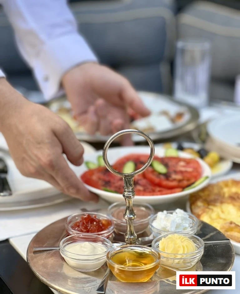 Gümüş renkli bir servis tepsisinde vişne reçeli, çikolata kreması, süzme bal, kaymak ve iki çeşit beyaz peynir/krema küçük cam kaselerde sunulmuşken, arka planda beyaz gömlekli bir kişinin masaya dilimlenmiş domates ve salatalık içeren bir tabak koyduğu görülüyor.