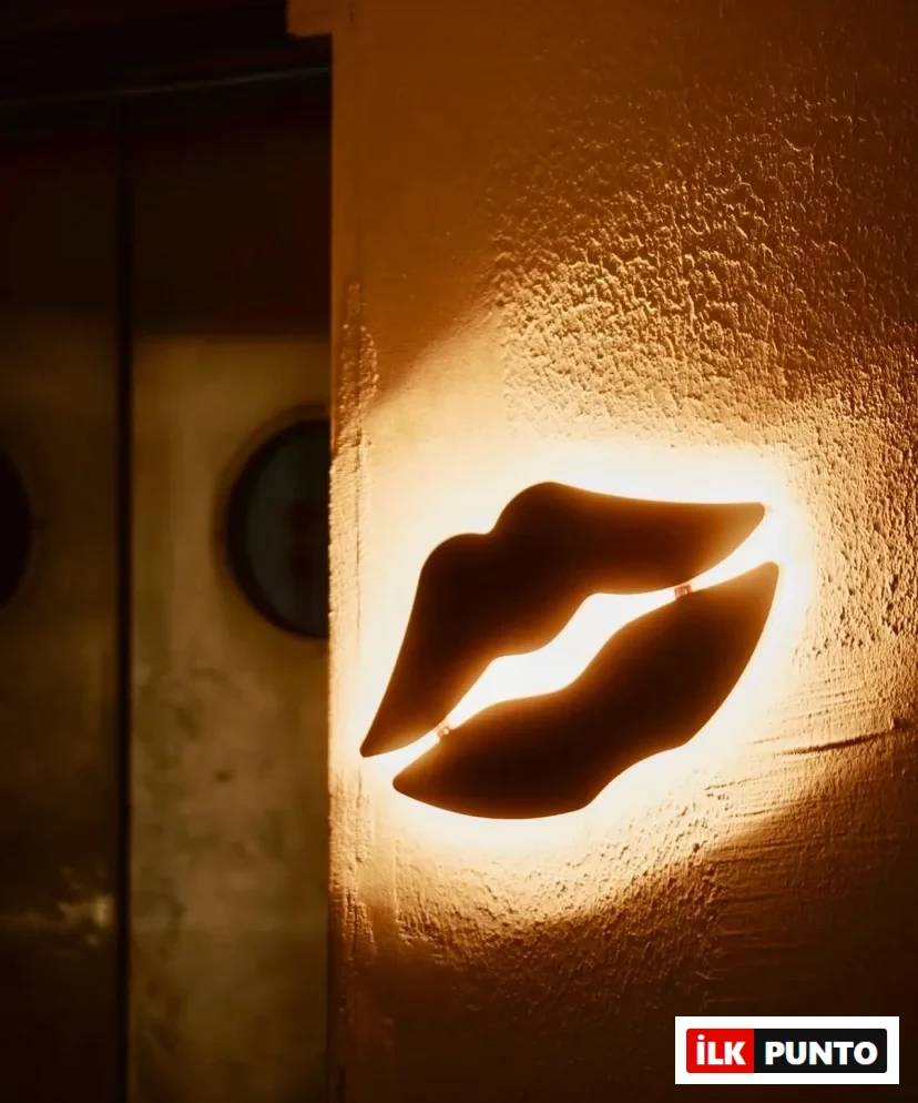 Dokulu ve sıcak sarı ışıkla aydınlanmış bir duvar üzerinde asılı duran, modern tasarımlı, parlak bir dudak figürü, arka planda belirgin olmayan dairesel detaylarla birlikte dikkat çekici ve farklı bir ambiyans yaratıyor.