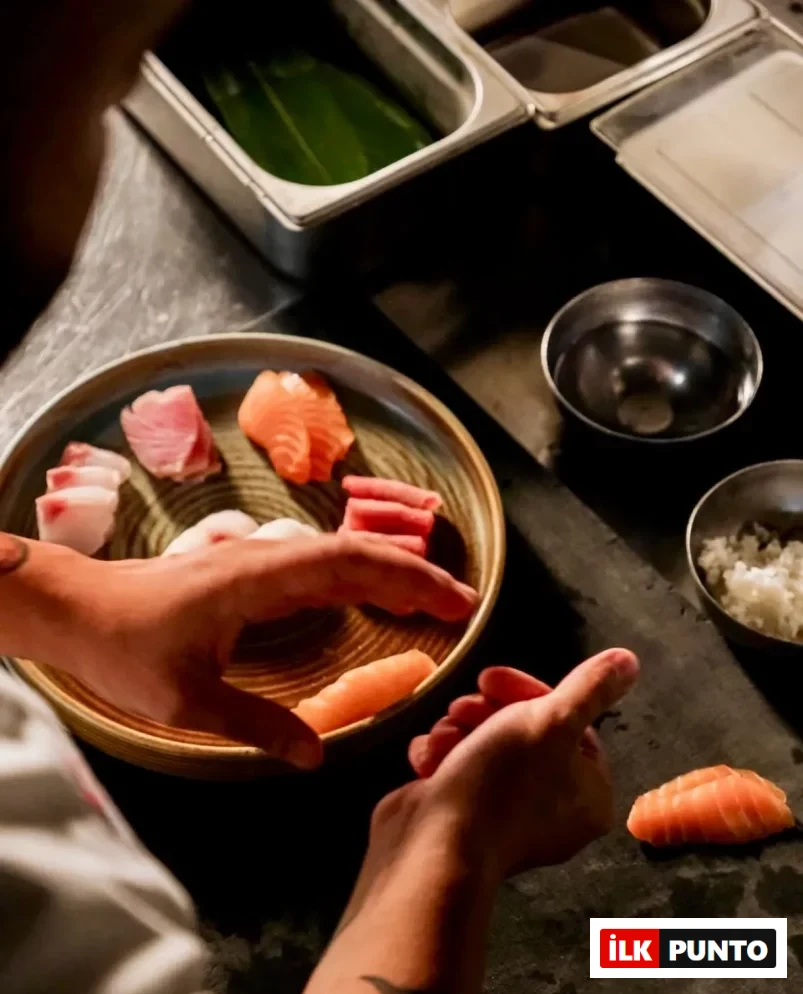 Bir şefin elleri, çeşitli somon, orkinos ve beyaz balık dilimleriyle dolu bir tabağın yanında, pirinci ustaca şekillendirerek özenle bir nigiri hazırlıyor; bu an, kişiselleştirilmiş bir omakase deneyiminin başlangıcını gözler önüne seriyor.