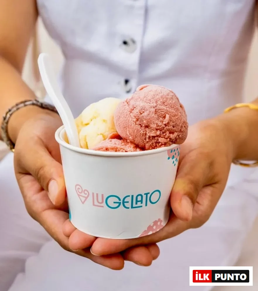 Beyaz kıyafetli bir kişinin ellerinde, üzerinde Lu GELATO logosu bulunan beyaz bir kap içinde, biri sarı diğeri pembe renkli iki top gelato ve beyaz bir kaşık görülüyor; bu serinletici lezzet, sıcak havalarda ferahlatıcı bir mola sunuyor.