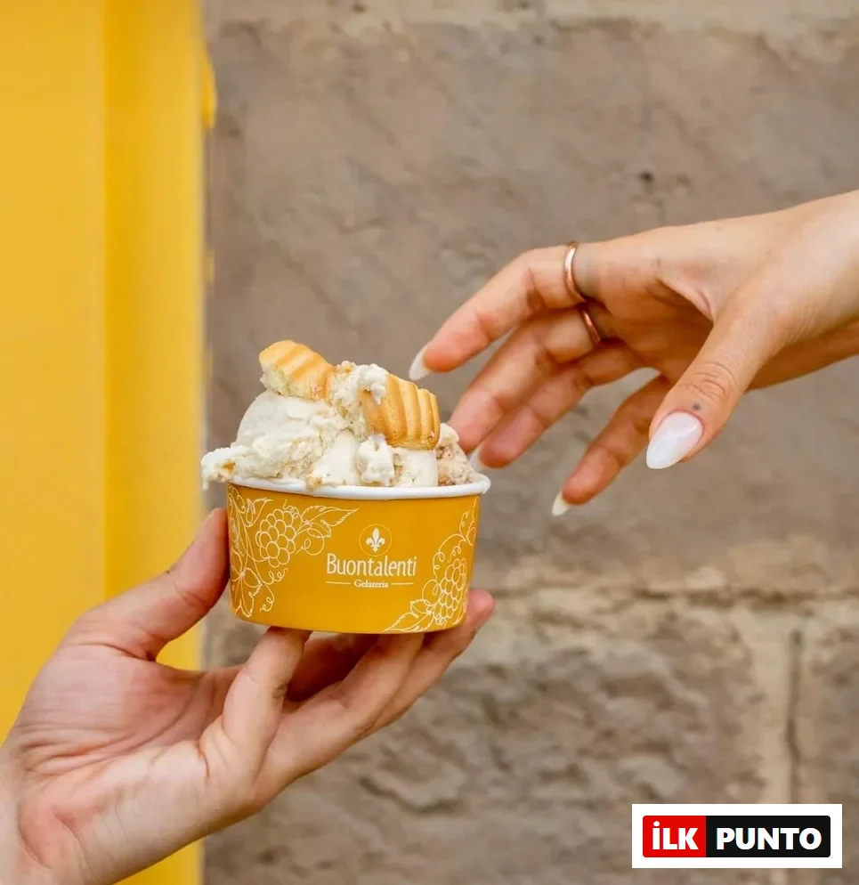 Sarı Buontalenti Gelateria kabında, üzerinde gofret veya kurabiye parçacıkları bulunan ve krem rengi bir gelato dururken, bir el onu tutuyor ve diğer el uzun beyaz tırnaklarıyla nazikçe kaba doğru uzanıyor.