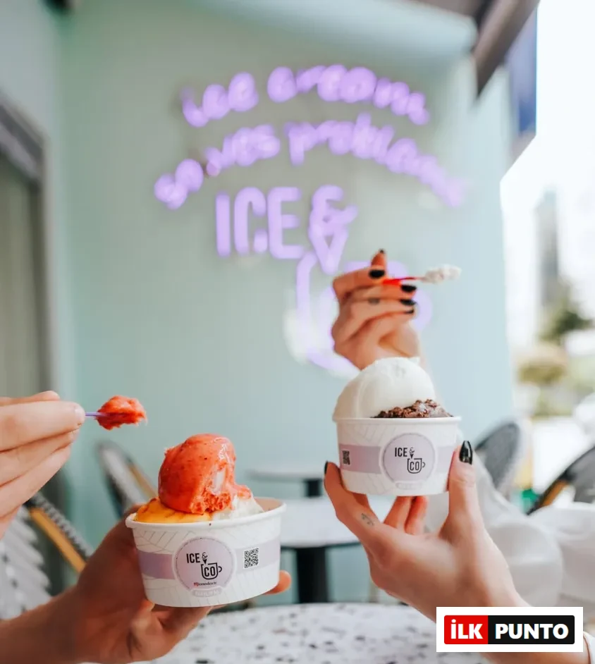 ICE & CO dondurma dükkanında iki el, çilekli ve portakallı bir dondurma ile çikolatalı ve vanilyalı başka bir dondurma kabı tutarken, üçüncü bir el kaşıklı dondurma servis ediyor; arka planda ise ICE & CO'nun neon tabelası dikkat çekiyor.