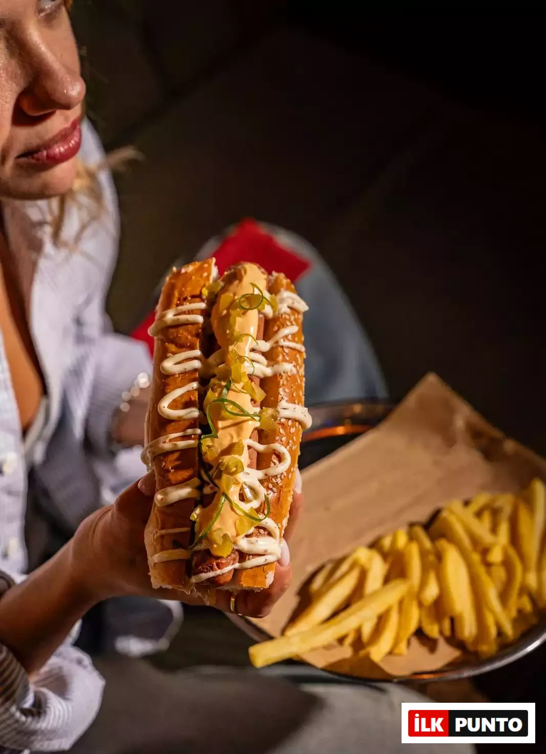 İstanbul’un: İlk Punto İstanbul'un En İyi Hamburgercileri: Bir kadın, elinde hardal, mayonez ve jalapeno biberleriyle süslenmiş, susamlı ekmek arası gurme bir sosisli sandviç tutuyor. Arka planda metal tepsi ve kraft kağıt üzerinde servis edilen çıtır patates kızartmaları iştah kabartıyor.