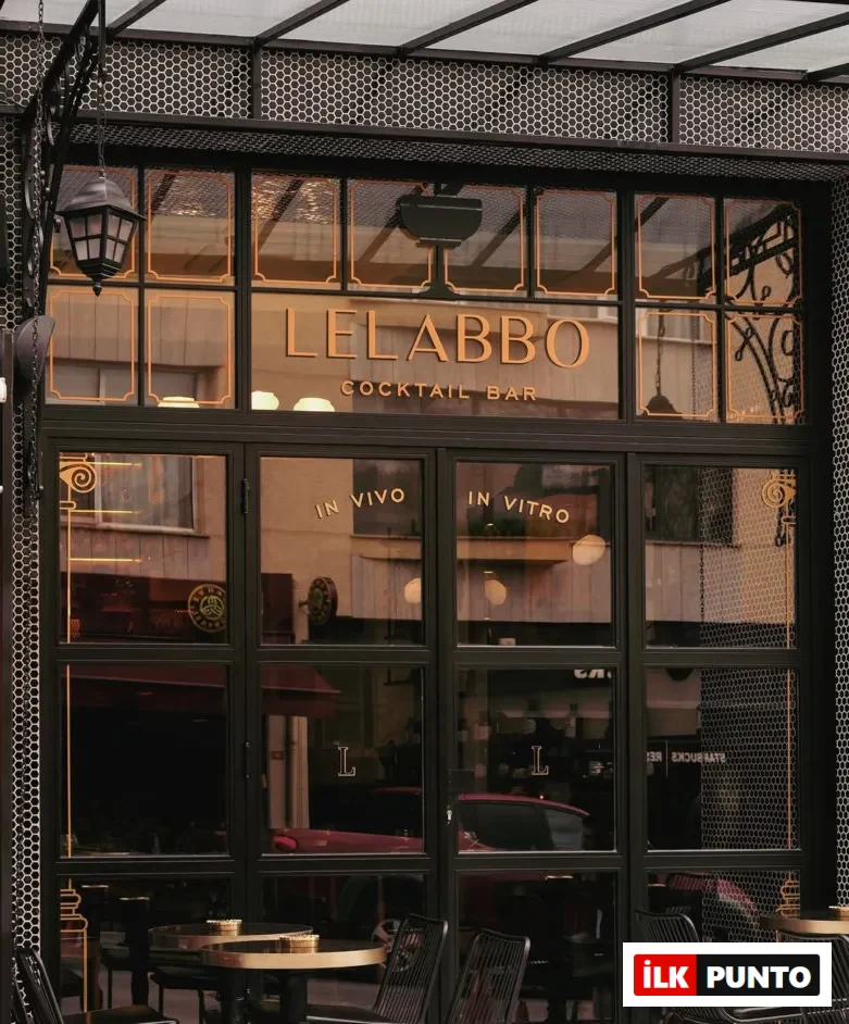Lelabbo Cocktail Bar'ın siyah çerçeveli cam cephesinde altın rengi LELABBO COCKTAIL BAR yazısı, IN VIVO ve IN VITRO ibareleri öne çıkarken, altıgen desenli kaplamaların çevrelediği girişin önünde altın tablalı yuvarlak masalar ve siyah sandalyeler zarif bir dış mekan oturma alanı sunuyor.