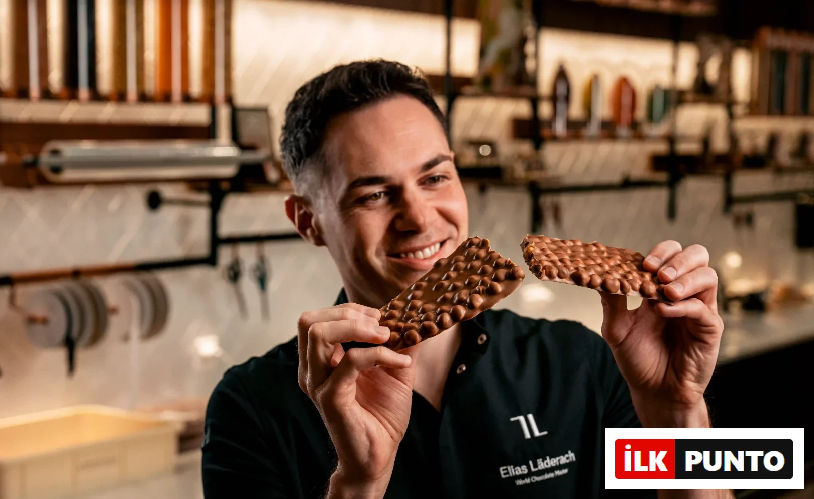 World Chocolate Master unvanlı Elias Läderach, elinde FrischSchoggi adı verilen, taze kırılmış, üzerinde bolca fındık bulunan bir sütlü çikolata parçasını gülümseyerek inceliyor; bu an, iyi çikolatanın zanaat ve tazelikle buluştuğunu gösteriyor.