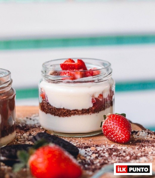İzmir'in en iyi pastanelerinden biri olan Mint Patisserie'de sunulan, ahşap bir sunum tahtası üzerinde yer alan, tabanı bisküvi kırıntıları, ortası beyaz krema ve üzeri bolca küp doğranmış taze çileklerle dolu, Magnolia tarzı cazip bir tatlı net bir şekilde görülüyor, etrafında taze çilekler ve bisküvi parçacıkları dağınık halde bulunuyor.