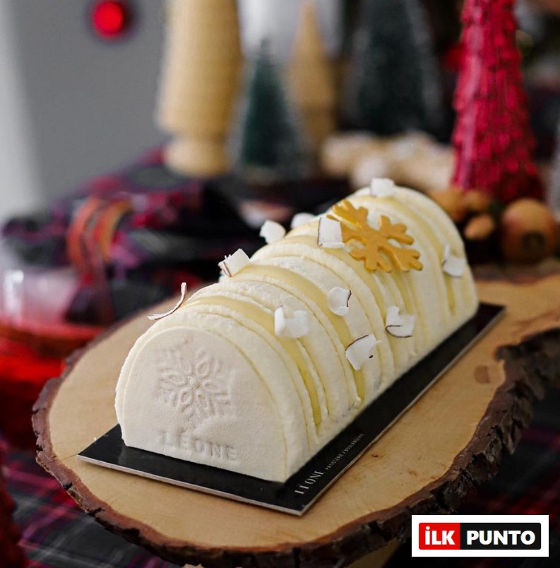 İzmir'in en iyi pastanelerinden Leone Patisserie'nin kış temalı sunumlarından, üzerinde zarif bir kar tanesi motifi ve marka adı işlenmiş, hindistan cevizi parçacıklarıyla süslü, dilimli, kütük şekilli beyaz pastası rustik bir ahşap servis tablasında sergileniyor.