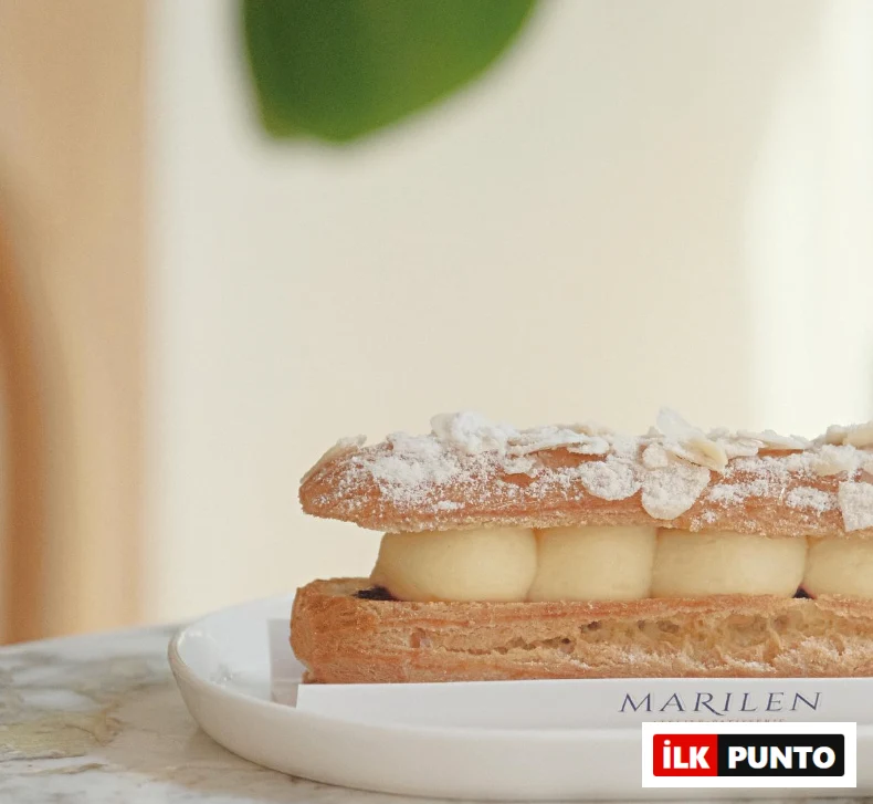 Beyaz bir tabakta, pudra şekeri ve badem dilimleriyle süslenmiş, pürüzsüz kremayla doldurulmuş, çıtır milföy tabanına sahip uzun bir Fransız pastası; İzmir'in en iyi pastanelerinden Marilen Atelier Patisserie'nin zarif sunumunu yansıtıyor.
