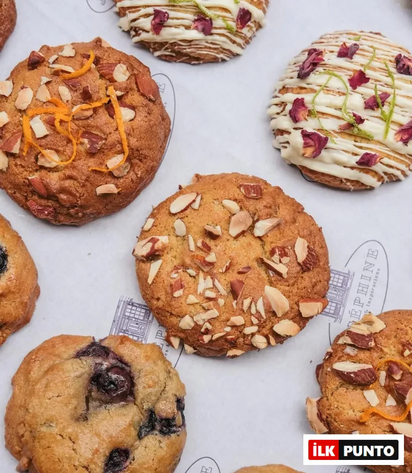 Joséphine Cookies'in farklı lezzetlerdeki kurabiyelerinin yakın çekim fotoğrafı. Kurabiyelerden bazılarında badem ve portakal kabuğu, bazılarında beyaz çikolata sosu ve gül yaprakları, bazılarında ise yaban mersini parçacıkları görülüyor.