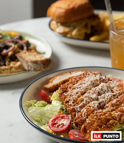 Suadiye'deki Nove Garden'da masada servis edilen, üzerinde rendelenmiş parmesan peyniri olan ve domates, marul ve mısırla hazırlanan tavuklu sezar salata. Arka planda hamburger ve patates kızartması da görünüyor.